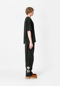 Camiseta negra de gran tamaño y pantalones a juego con una superficie texturizada. Los pantalones presentan un bordado de un oso gris en el dobladillo. Zapatos marrones con suelas gruesas.