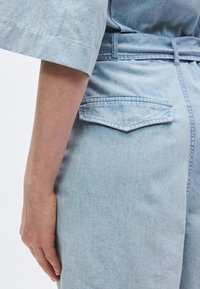 Ελαφρώς γαλάζιο ρούχο από τζιν με χαλαρή εφαρμογή, φαρδιά μανίκια και ζώνη στη μέση. Διακριτική πίσω τσέπη με στρογγυλεμένο καπάκι.