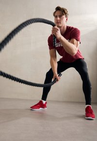 Sort kampesejl holdt af en fit person i en rød t-shirt og sorte leggings, der udfører en øvelse på et glat, lys gråt gulv.