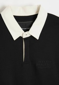 Czarna koszulka polo z kremowym kołnierzykiem, zapięciem na guziki oraz haftowanym logo „Różnorodne Dziedzictwo” po lewej stronie. Miękka, żebrowana tekstura.
