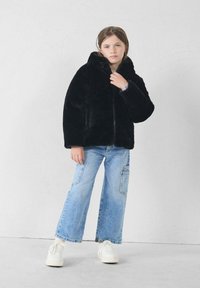 Chaqueta de piel sintética negra con capucha, mangas largas y cremallera frontal, combinada con vaqueros de pierna ancha azul claro y zapatillas blancas.