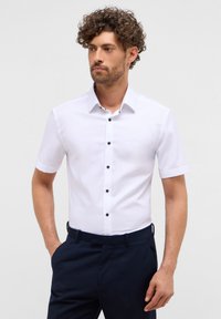 Camicetta bianca a maniche corte con bottoni neri e colletto classico, texture liscia, abbinata a pantaloni blu navy.