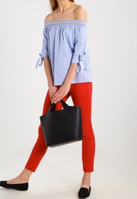 Hellblaues Off-Shoulder-Bluse mit gerafftem Ausschnitt, Dreiviertelärmeln und gebundenen Manschetten. Kombiniert mit engen roten Hosen und einer schwarzen Tragetasche.