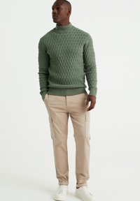 Zeleni pleteni pulover s turtleneck okraskom in teksturirano diamantno vzorcem, v kombinaciji s bež cargo hlačami, ki imajo stranske žepe, ter belimi supergami.