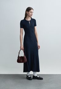 Kvinne i marineblå knyttet knappet strikkekjole som holder en burgunder håndveske, iført hvite sokker og svarte Mary Jane-sko, står på en grå gulvflate.