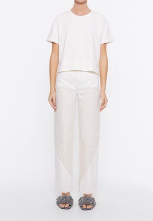 T-shirt bianco a maniche corte con collo rotondo, abbinato a pantaloni larghi beige chiaro con dettagli di cuciture diagonali. Sandali con zeppa grigi a fiori.