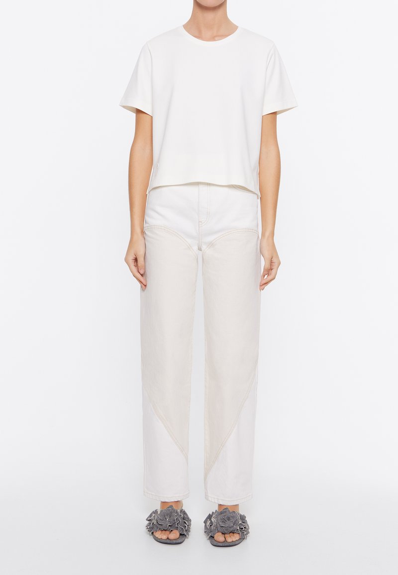 T-shirt bianco a maniche corte con collo rotondo, abbinato a pantaloni larghi beige chiaro con dettagli di cuciture diagonali. Sandali con zeppa grigi a fiori.