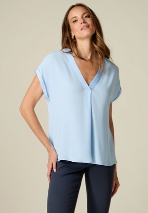 Vrouw draagt een lichtblauwe korte mouwen V-hals blouse en donkere broek, poseert met één hand op haar heup tegen een beige achtergrond.