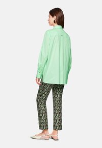 Helles grünes, übergroßes Hemd mit Knopfmanschetten, kombiniert mit schwarzen und grünen Hosen im Blätterdesign und weißen Slingback-Schuhen.