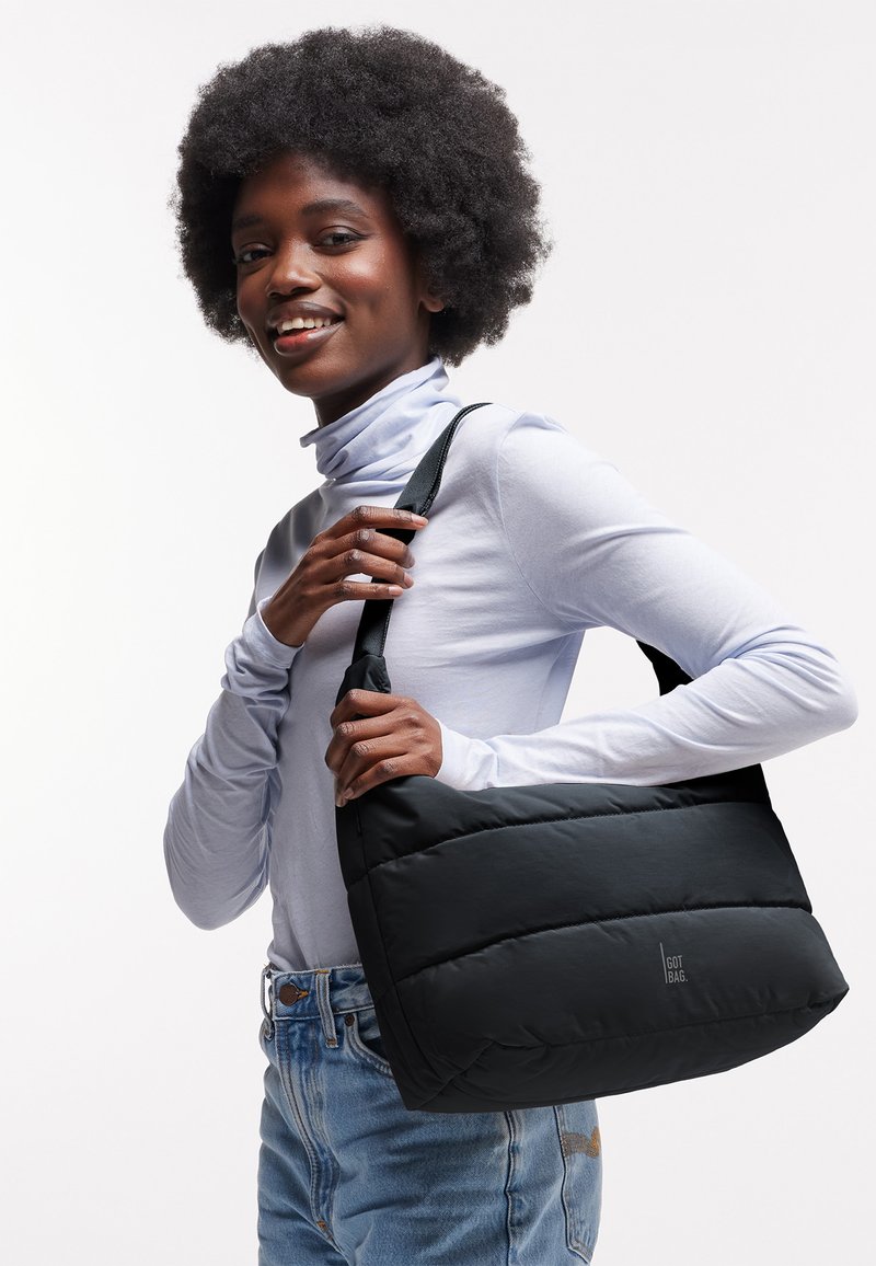 Kvinde med afrofrisure iført en hvid polo og jeans holder en sort polstret skuldertaske med teksten "GOT BAG".