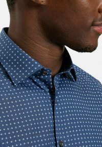 Marineblaues Hemd mit kleinem weißem und hellblauem Blumenmuster, mit spitzem Kragen und schwarzem Knopf. Glatte Stoffstruktur.