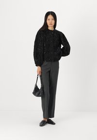 Schwarze, strukturierte Bluse mit Puffärmeln, kombiniert mit grauen Streifenhose und schwarzen Slippers, hält eine kleine schwarze Handtasche.