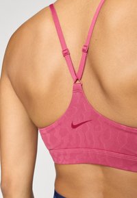 Vaaleanpunainen urheiluliivi, jossa on leopardikuosiset yksityiskohdat, säädettävät hihnat ja pieni marooni logo. Liivissä on sileä pinta ja tiukka muotoilu.