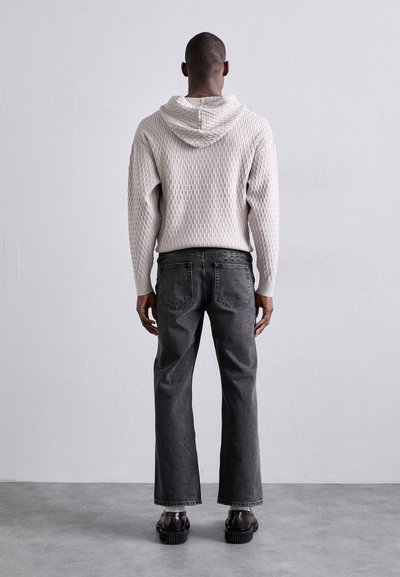 Haut à capuche en tricot gris clair avec un motif texturé, associé à un jean large gris foncé et des chaussures noires épaisses, vu de dos.