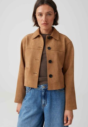Donna che indossa una giacca beige con bottoni sopra una maglietta marrone e jeans blu, in piedi contro uno sfondo grigio chiaro uniforme.
