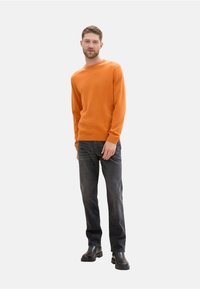 Suéter de punto color naranja, con cuello redondo; combinado con jeans gris oscuro. La modelo lleva botas negras hasta el tobillo. Diseño simple, tejido texturizado.