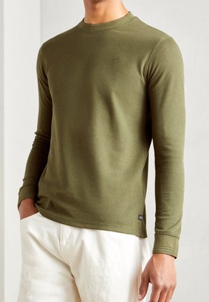 Man draagt een olijfgroen textuur lang shirt met lange mouwen en witte broek, staand met één hand in de zak tegen een effen achtergrond.