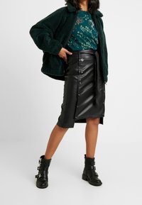 Jupe en cuir noir avec fermeture à boutons sur le devant et fente sur le côté, associée à une veste texturée verte et un haut à imprimé floral. Bottines noires.