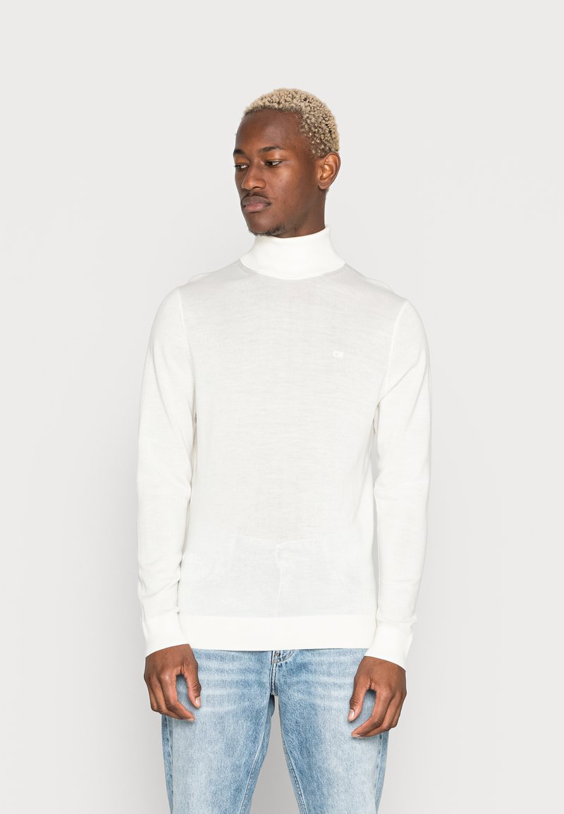 Hvid, langærmet turtleneck sweater med en fin strikstruktur og et lille logo på brystet, parret med lyseblå jeans.