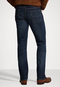 Muž v tmavě modrých slim-fit džínách a hnědých kožených botách, viděn zezadu na jednobarevném světlém pozadí.