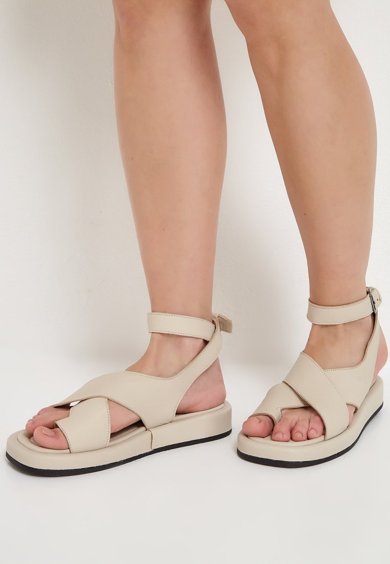 Beige sandaler med korsade remmar, ankelsnodd och platt sula. Tillverkade av slätt material med rund tå och texturerade detaljer.
