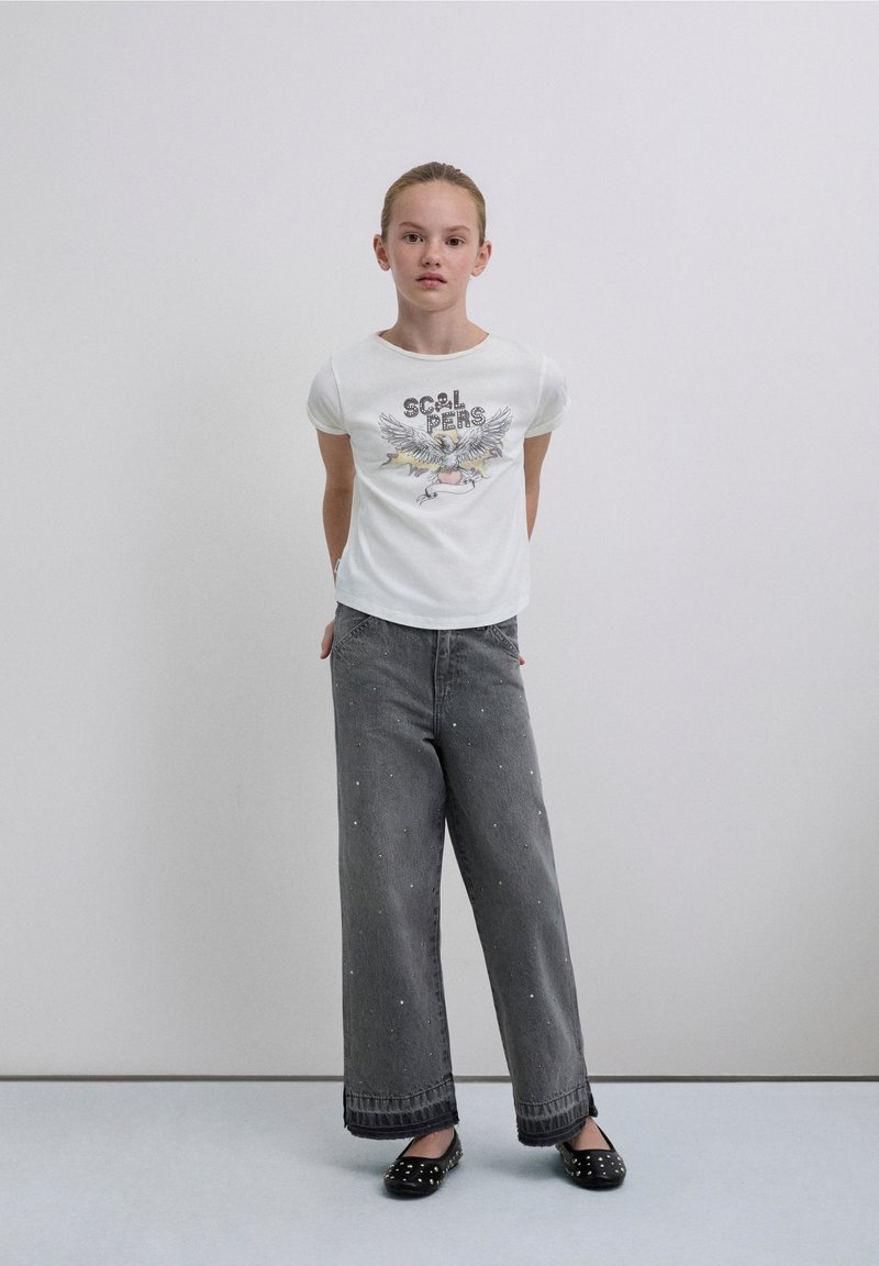 T-shirt bianca in cotone con stampa grafica, abbinata a jeans grigi a gamba larga in denim impreziositi da piccole borchie. Scarpe piatte nere.