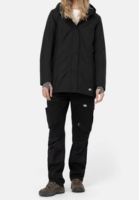 Svart vattentät jacka med huva, fleecefoder; kombinerad med svarta cargo-byxor med fickor och förstärkta knän; bärs med bruna vandringsskor.