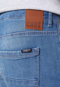 Pantalones vaqueros de mezclilla azul desgastados con una etiqueta de cuero marr ón que dice "CARS" y una etiqueta negra con la inscripción "CARS™" en el bolsillo trasero.