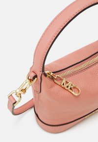 Bolso de piel rosa con herrajes dorados, forma redondeada, asa superior y cierre de cremallera que presenta un charm de "MK" y un ribete negro contrastante.