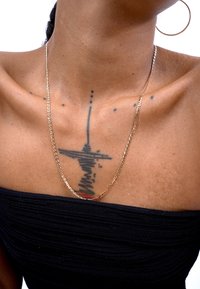 Gold-colored chain necklace resting on bare skin near a tattoo. The necklace has a flat, thin design with a textured surface.