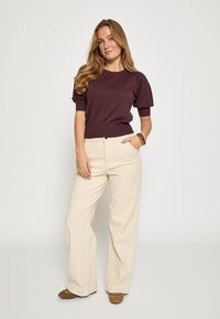 Vínová krátká pletená svetr s ribbovaným lemem, doplněný krémovými kalhotami s vysokým pasem a širokými nohavicemi. Hnědé boty dotvářejí celý outfit.