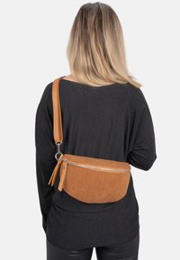 Braune Korduratasche mit einem Reißverschluss, langem verstellbarem Tragegurt und einer subtilen Rillenstruktur. Am Rücken einer Person getragen.