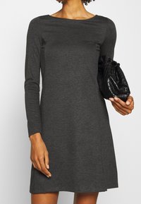 Grå långärmad klänning med båturringning, tillverkad av slät tyg. I kombination med en svart strukturerad handväska. Enkel A-linjeform.