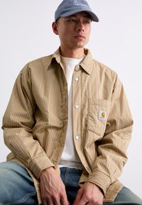 Striped beige shirt with white accents, button-up front, and chest pocket. Worn over a white t-shirt and paired with blue jeans.