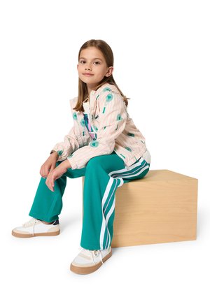 Jong meisje zittend op een houten blok, gekleed in een roze jas met patroon, groene trainingsbroek met witte strepen en witte sneakers.