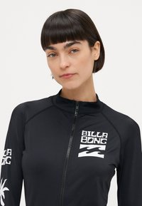 Črni zip-up rashguard z dolgimi rokavi, ki ima bel logo "BILLA BONG" in palmni grafik na rokavih, gladka tekstura, zmogljiv tkanina.