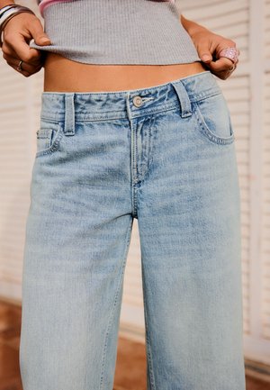 Ruky ťahajú svetlosivý rebrovaný tielko nahor, odhaľujúc bruško nad svetlomodrými džínami s vysokým pásom s gombíkom a vreckami.