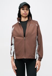 Καφέ ανοιξιάτικο μπουφάν με γκρι λεπτομέρειες, ολόσωμο φερμουάρ, ελαστικές μανσέτες και ψηλό γιακά. Λογότυπο στο αριστερό στήθος, συνδυασμένο με μαύρο καπέλο.