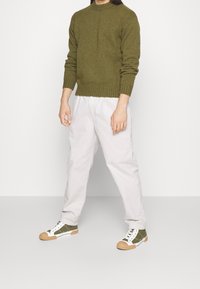 Person som bär en olivgrön tröja, ljusbeiga byxor och olivgröna sneakers med vita snören, står mot en enkel ljus bakgrund.