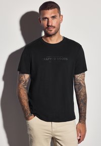 Muž s tetováním, oblečený v černém tričku "Crafted Goods" a béžových kalhotách, stojící proti bílé zdi a vrhající stín.