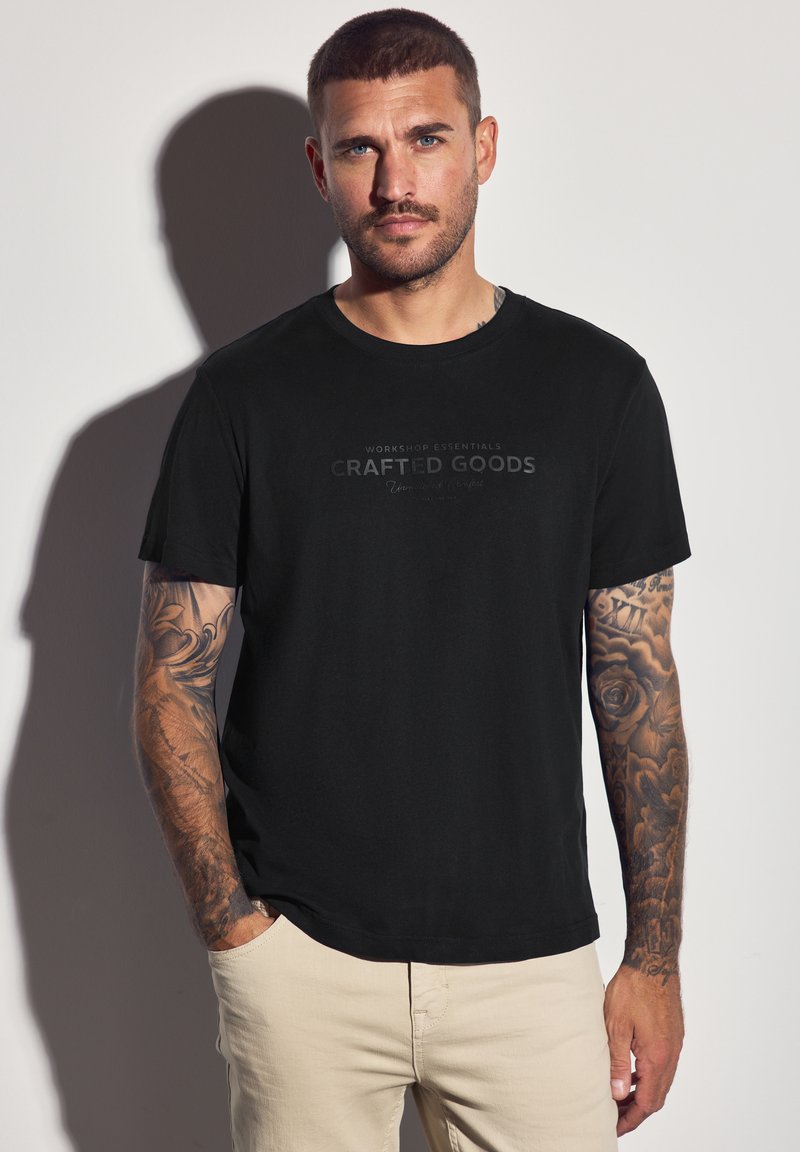 Muž s tetováním, oblečený v černém tričku "Crafted Goods" a béžových kalhotách, stojící proti bílé zdi a vrhající stín.