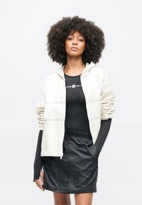 Φωτεινό μπεζ μπουφάν με φερμουάρ, μακριά μανίκια και μπροστινές τσέπες, στρωμένο πάνω από μαύρο εφαρμοστό τοπ και μαύρη φούστα με ελαστική μέση. Λιτή υφή.