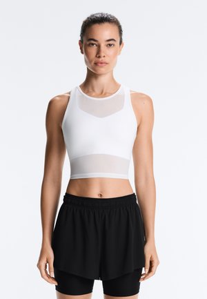 Kvinde står iført hvid ærmeløs cropped top og sorte lagdelte atletikshorts med tætsiddende leggings under mod en ensfarvet baggrund.