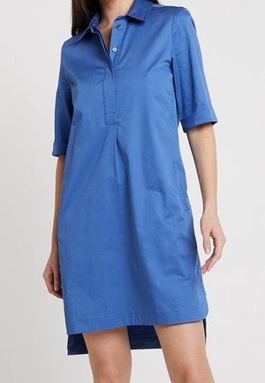 Niebieska sukienka-koszula do kolan z kołnierzykiem, półdługimi rękawami, guzikami z przodu i bocznymi kieszeniami, noszona przez stojącą osobę.