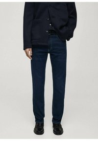 Tamno plave traperice od denima, ravnog kroja s lagano izblijeđenim završnim obradama. Uparene s crnom, dugmad-up vanjskom odjećom. Crne cipele.