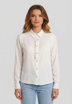 Donna con capelli ondulati di media lunghezza indossa una camicetta bianca con bottoni e dettagli con borchie e jeans azzurri, in piedi davanti a uno sfondo grigio.