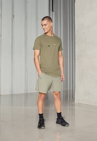 Olive green short-sleeve t-shirt with a textured, black Nike logo; light green shorts; black athletic shoes; smooth floor, metallic backdrop.