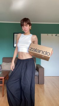 Bílé bezrukávové crop top a černé široké kalhoty. Drží hnědou krabici Zalando. Místnost se zelenými stěnami a světlou dřevěnou podlahou.