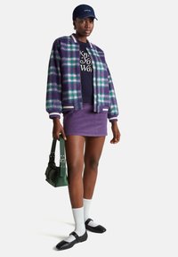 Chaqueta bomber de cuadros púrpuras con rayas blancas, camiseta negra, falda corta púrpura, calcetines blancos, zapatos negros y bolso verde.