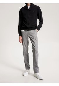 Man som bär en svart zip-up-tröja, vit skjorta, grå byxor och vita sneakers, stående med en hand i fickan mot en enfärgad bakgrund.